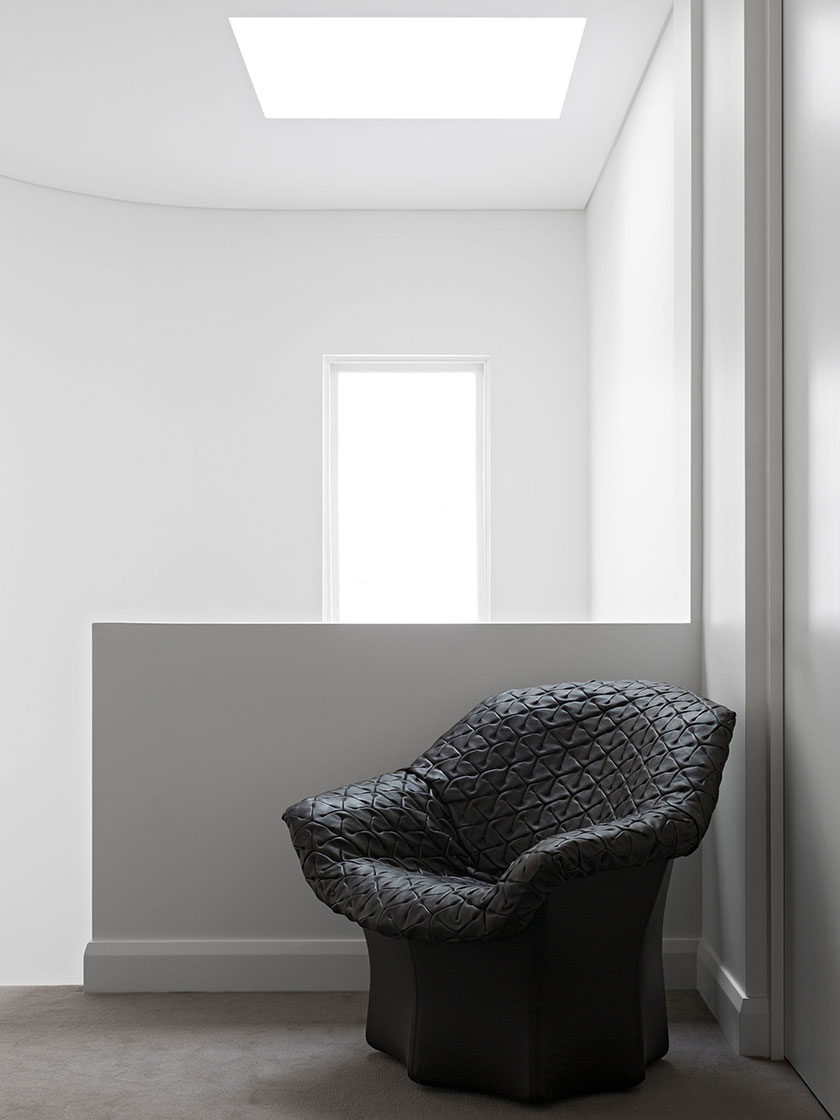 Minimalist hallway with a black chair, white walls, and a skylight, designed by Nina Maya Interiors.