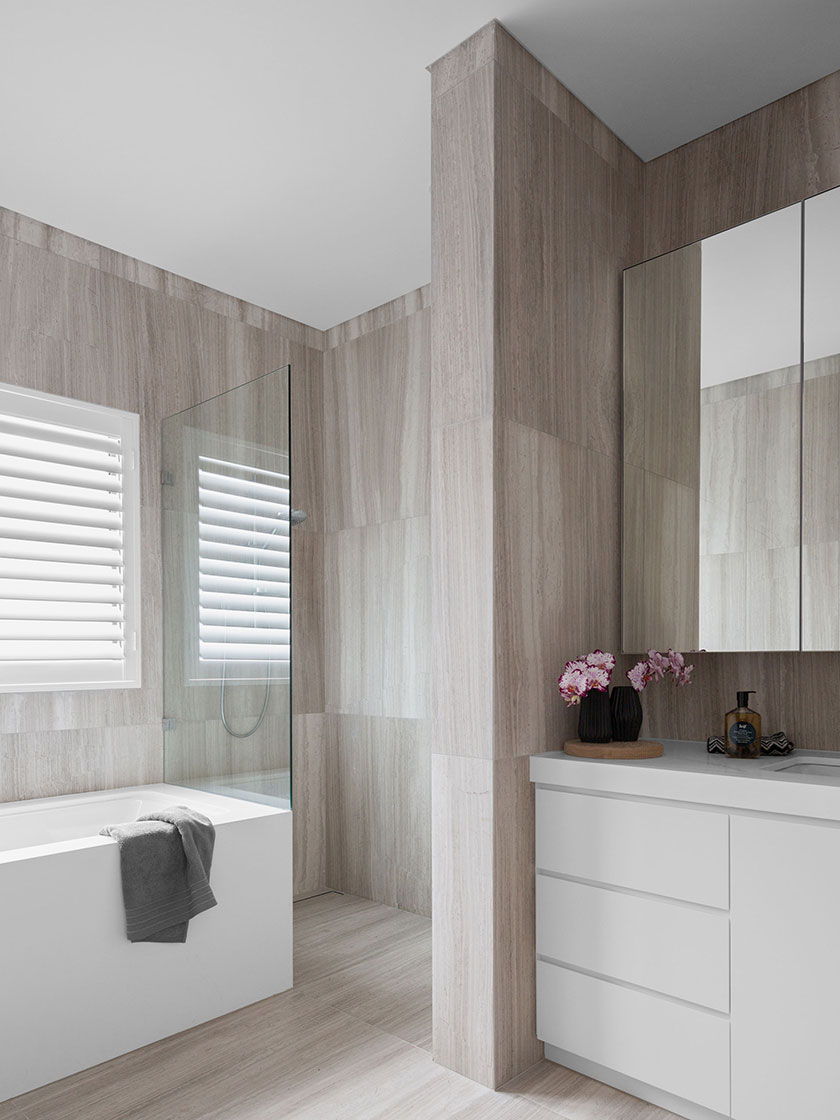 Modern bathroom with beige textured tiles, white bathtub, and vanity with pink flowers, designed by Nina Maya Interiors.