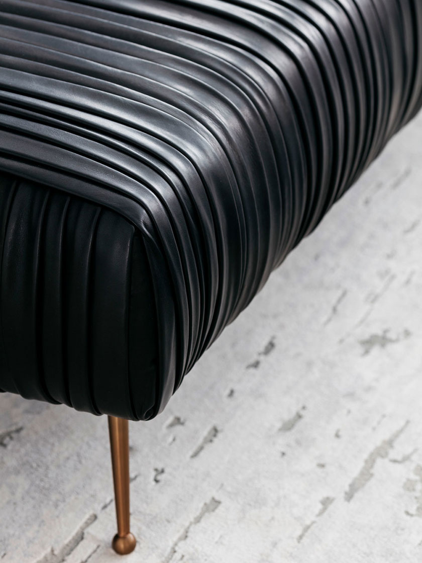 Close-up view of a black leather ottoman with brass legs on a light grey rug.