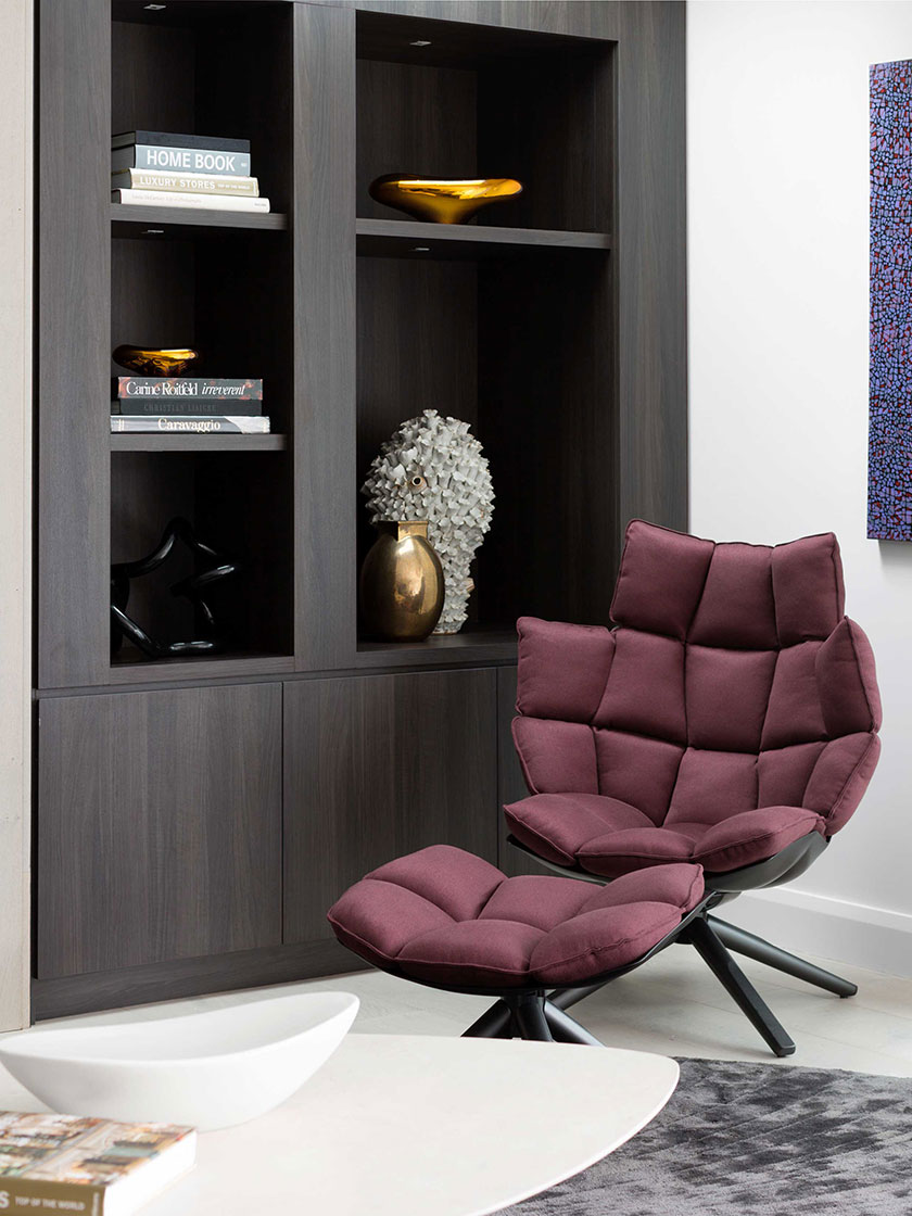 Stylish reading corner with a burgundy lounge chair and ottoman, built-in dark wood bookshelf, and modern decor, designed by Nina Maya Interiors.